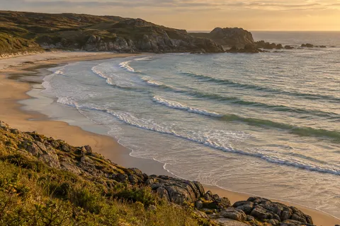 Gentle surf arcs around a rocky sandy bay in warm evening light.