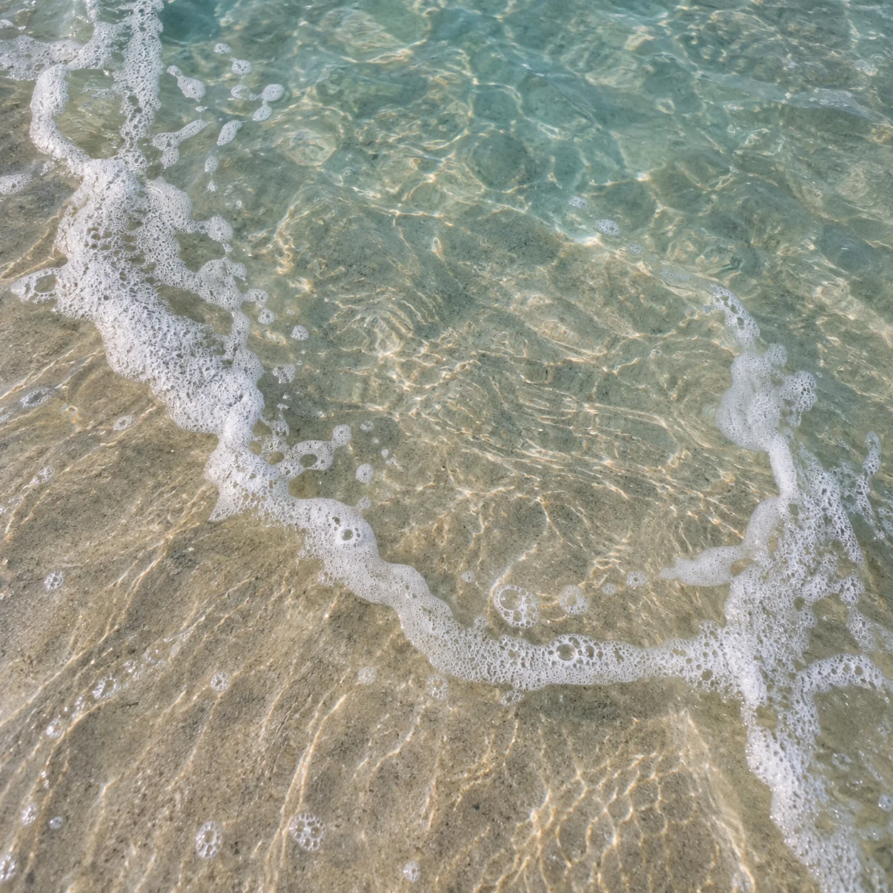 A thin curling foam trail crosses clear shallow water above pale sand.