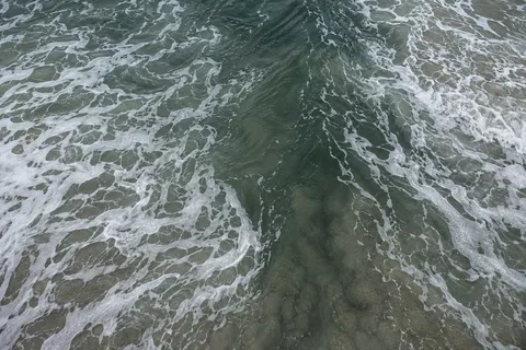 Retreating foam opens around a dark channel in shallow gray-green water.