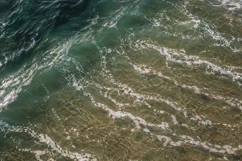 Thin white foam strands drift over green and bronze shallows beside a dark diagonal band of water.