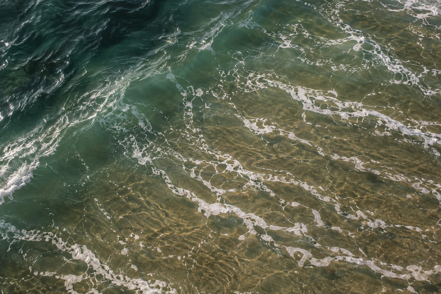Thin white foam strands drift over green and bronze shallows beside a dark diagonal band of water.