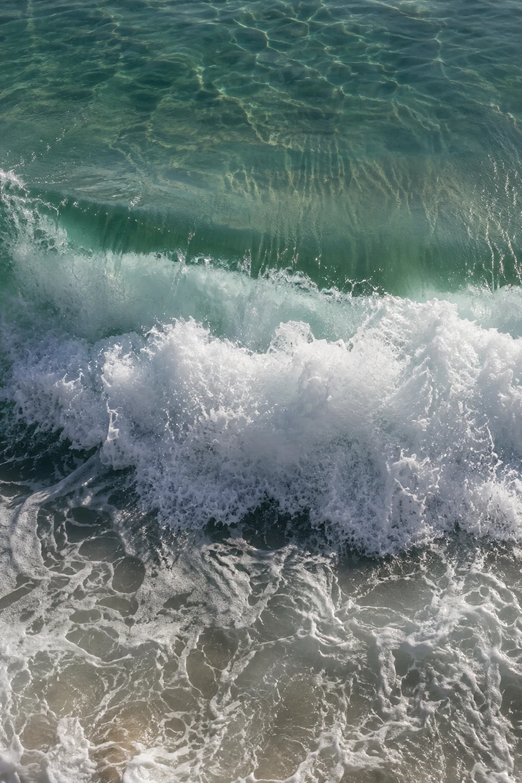 A bright breaking wave crosses clear green shallows above pale submerged sand.