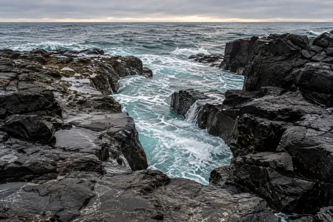 Pale turquoise surf enters a jagged opening between wet black coastal rocks beneath a gray sky.
