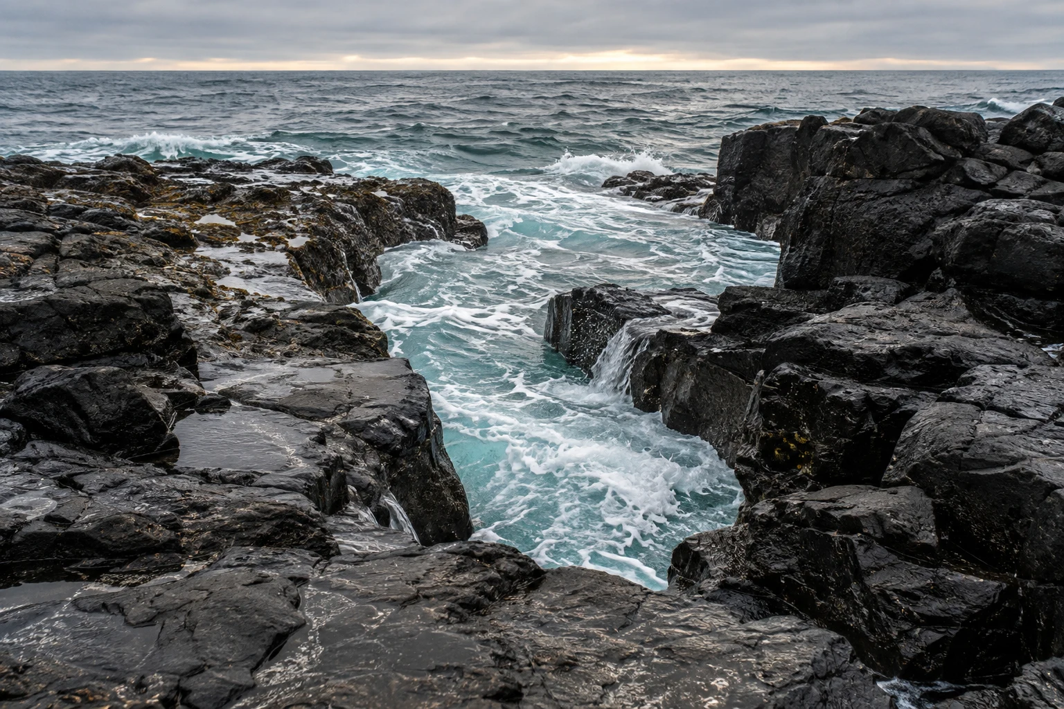 Pale turquoise surf enters a jagged opening between wet black coastal rocks beneath a gray sky.