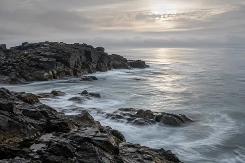 Dark rock shelves extend into a silver sea beneath softly lit cloud.