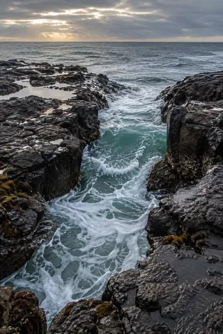 Green water surges through a narrow volcanic channel below a break in the clouds.