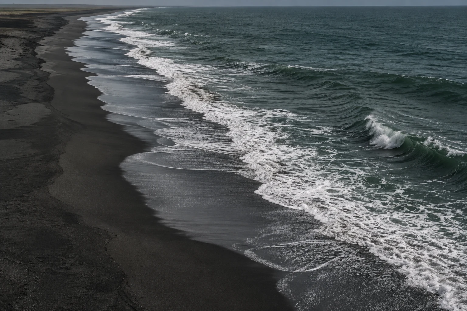 A winding white shorebreak traces a long empty black-sand coast.