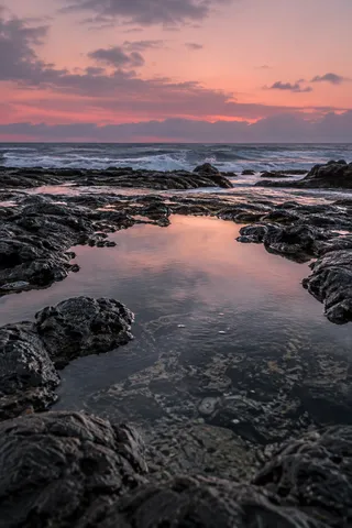 A shallow pool in coarse black rock reflects a rose-colored evening sky beyond the sea.