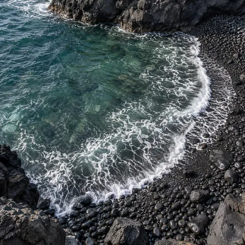 A curved wave laps a small cove of smooth black pebbles beside clear green water.