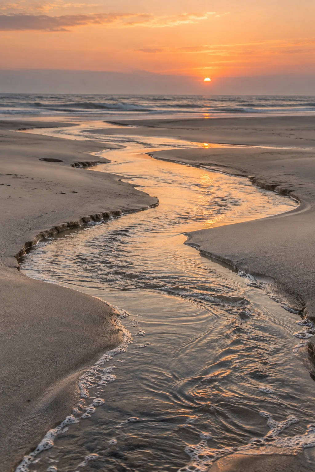 A winding shallow channel catches orange sky light on its way through sand to the sea.