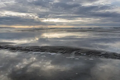 Clouds reflect across broad tidal shallows separated by a narrow rippled sandbar.