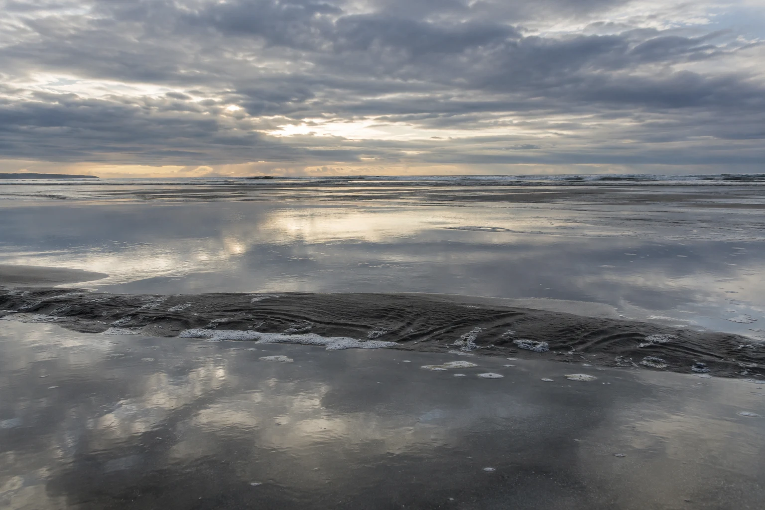 Clouds reflect across broad tidal shallows separated by a narrow rippled sandbar.