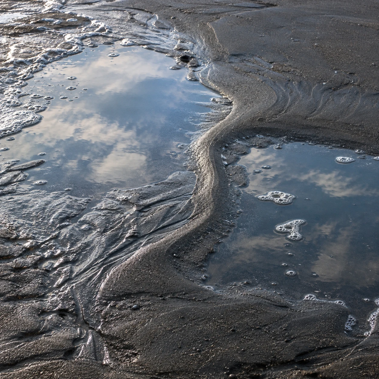 Two shallow pools reflect clouds beside a winding ridge of dark wet sand.