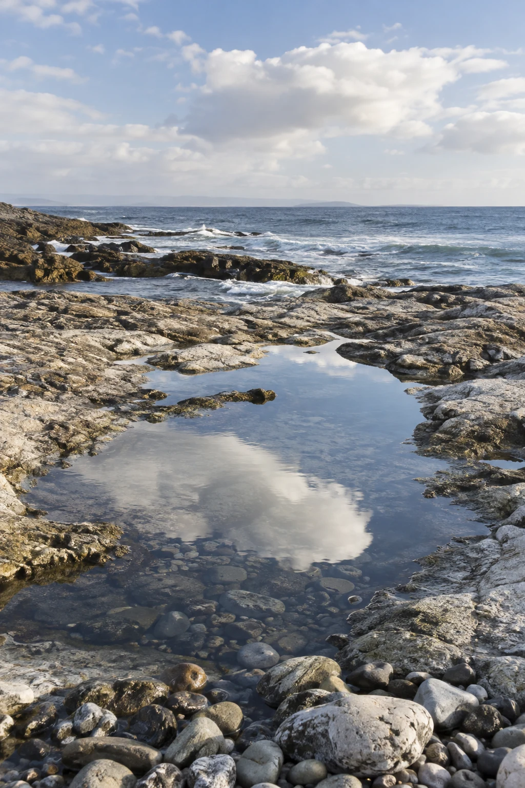 A clear pool between pale coastal rocks reflects a passing cloud above submerged stones.