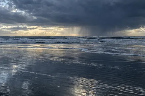 A dark rain cloud hangs over the sea while pale light reflects across wet tidal sand.