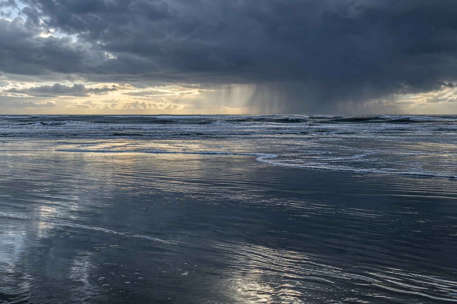 A dark rain cloud hangs over the sea while pale light reflects across wet tidal sand.