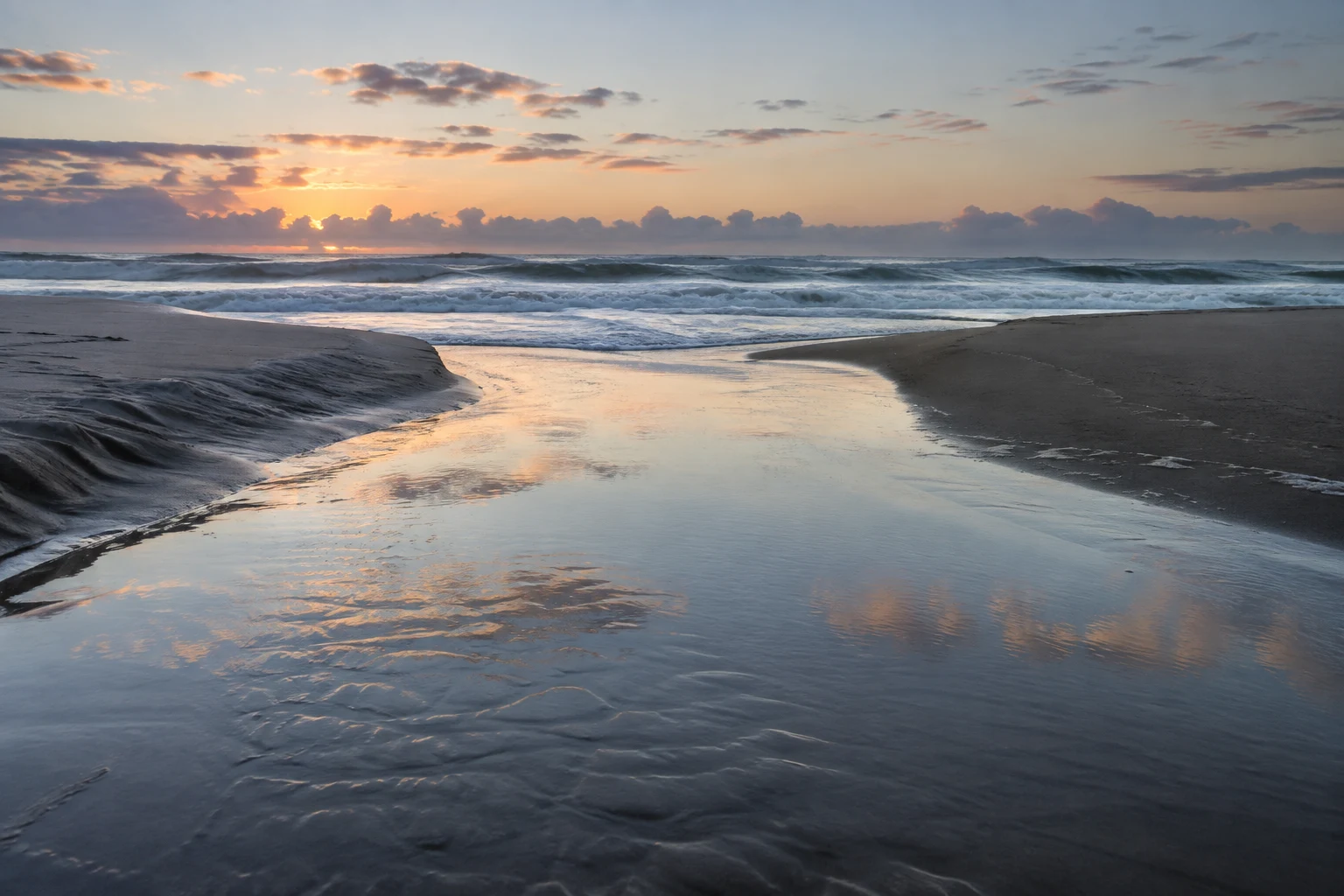 A quiet tidal channel opens between dark sandbanks toward breaking waves under a pale evening sky.