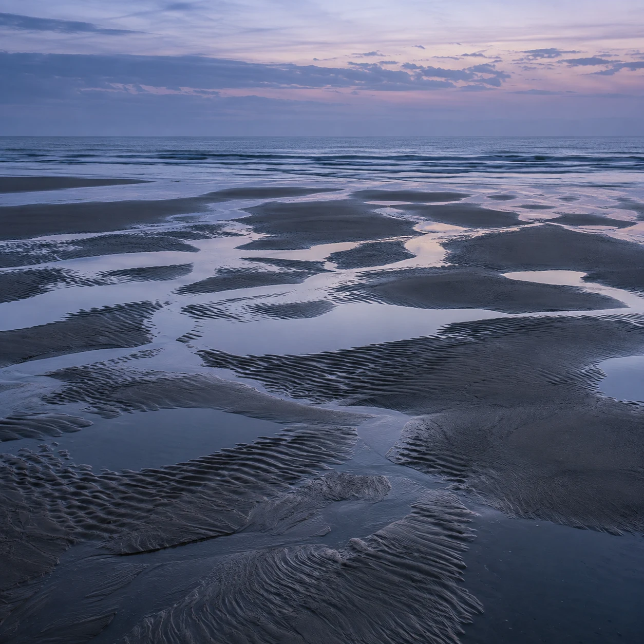 Shallow channels weave among rippled sand flats under a soft blue evening sky.