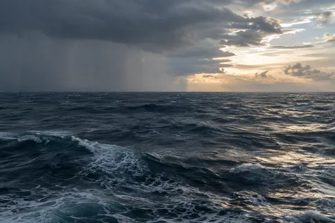 A rain front crosses dark blue sea beside a warm opening in the clouds.