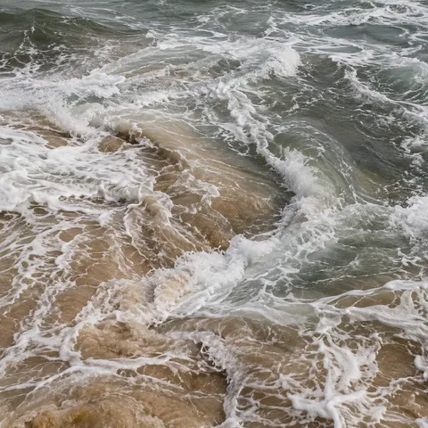 Foamy currents meet over sandy shallows in a close view of gray-green surf.