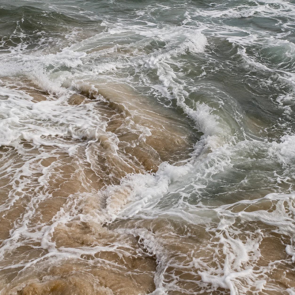 Foamy currents meet over sandy shallows in a close view of gray-green surf.