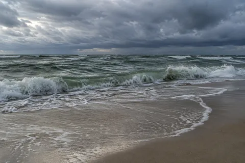 A green-gray wave breaks along pale sand under a heavy band of cloud.