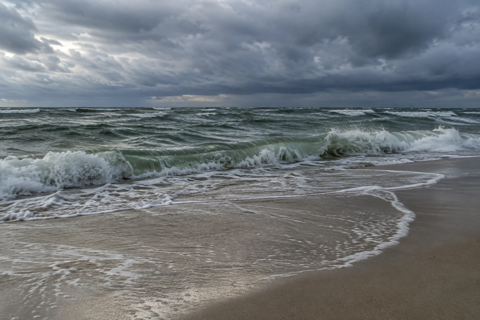 A green-gray wave breaks along pale sand under a heavy band of cloud.