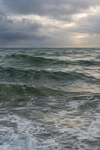 Soft light returns through gray clouds above green swells and nearshore foam.