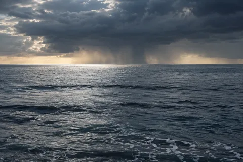 A distant rain curtain hangs below dark clouds over a broad blue-gray sea.