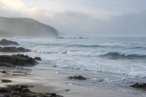A green headland fades into pale sea mist above scattered dark rocks and low white waves.