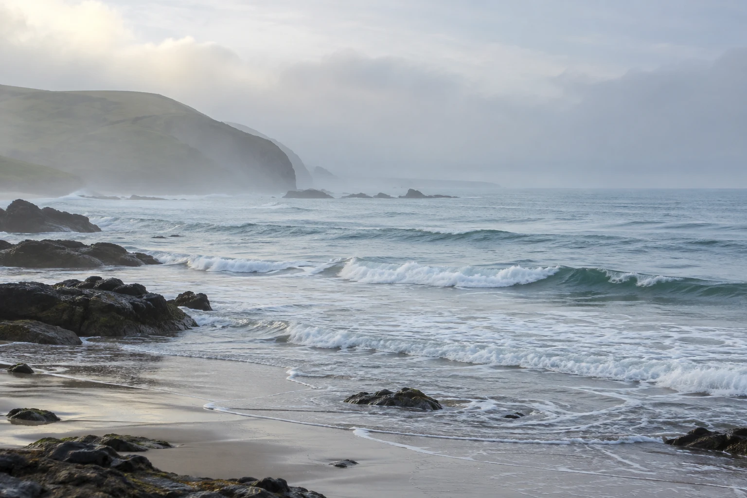 A green headland fades into pale sea mist above scattered dark rocks and low white waves.
