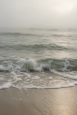 A small gray-green wave breaks toward a sandy shore beneath a broad field of pale fog.