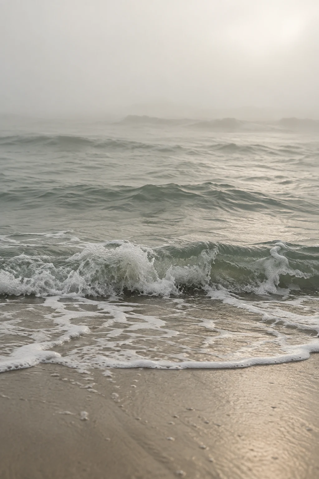 A small gray-green wave breaks toward a sandy shore beneath a broad field of pale fog.