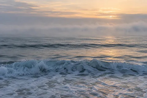 Low blue-gray waves cross a calm sea beneath a horizontal fog bank and a pale warm sky.
