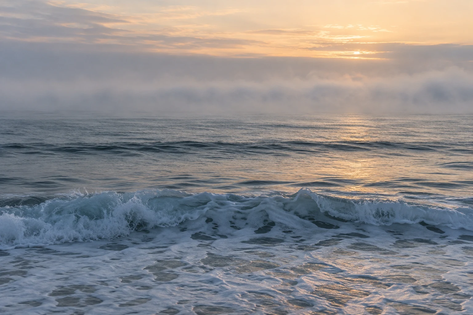 Low blue-gray waves cross a calm sea beneath a horizontal fog bank and a pale warm sky.