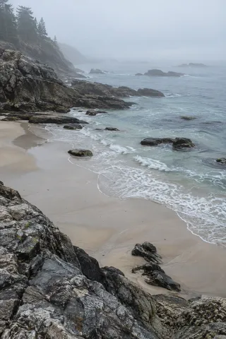 Dark foreground rocks frame a sandy cove whose distant shoreline dissolves into white mist.