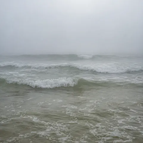 Small gray-green waves form pale parallel bands beneath an almost white foggy distance.