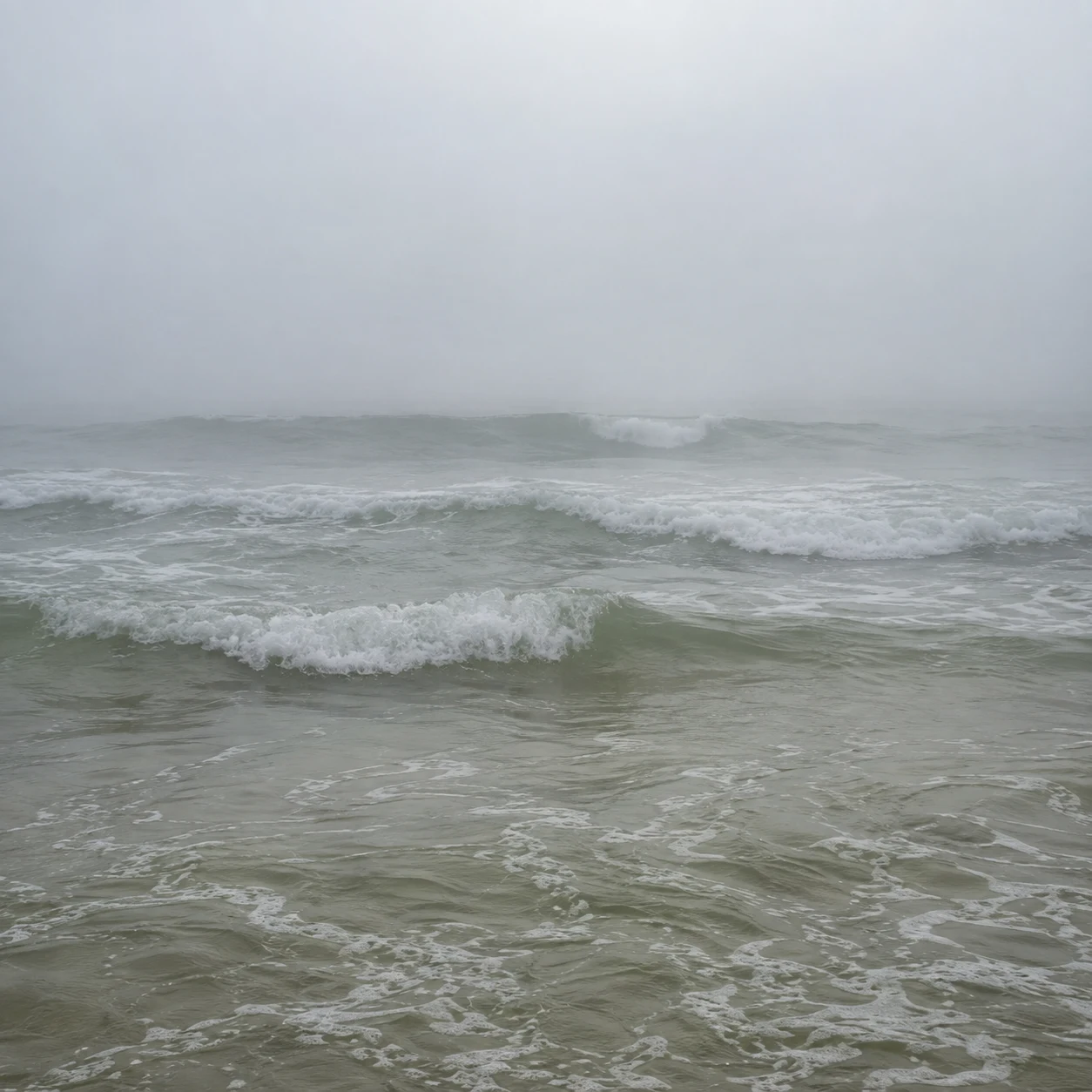 Small gray-green waves form pale parallel bands beneath an almost white foggy distance.