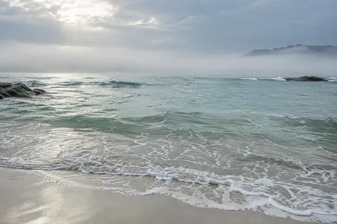 Fine surf crosses pale sand below a milky blue-green bay and a distant misty wooded point.