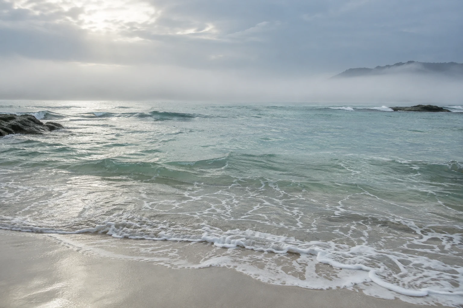 Fine surf crosses pale sand below a milky blue-green bay and a distant misty wooded point.