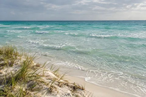 Sparse dune grass borders pale sand beside a wide expanse of clear turquoise shallows.