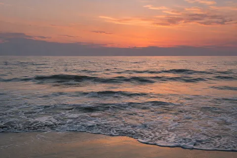 Peach and rose clouds hang above low slate-blue waves and a reflective wet shoreline.