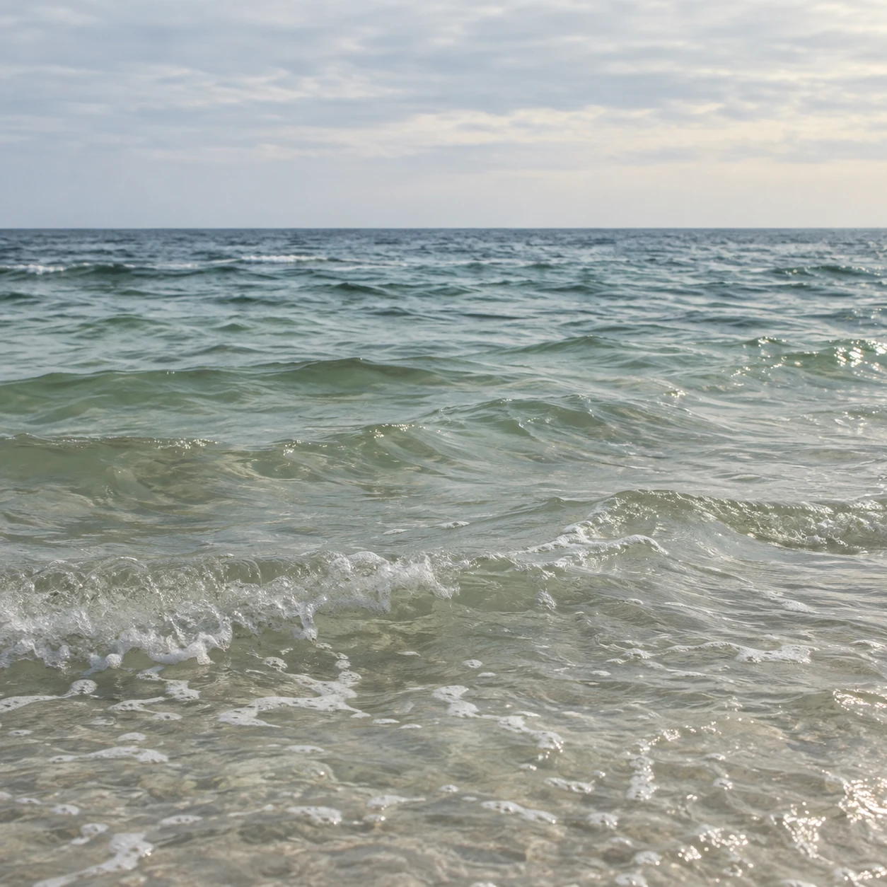 Clear pale green wavelets ripple toward a distant blue-gray horizon under thin cloud.