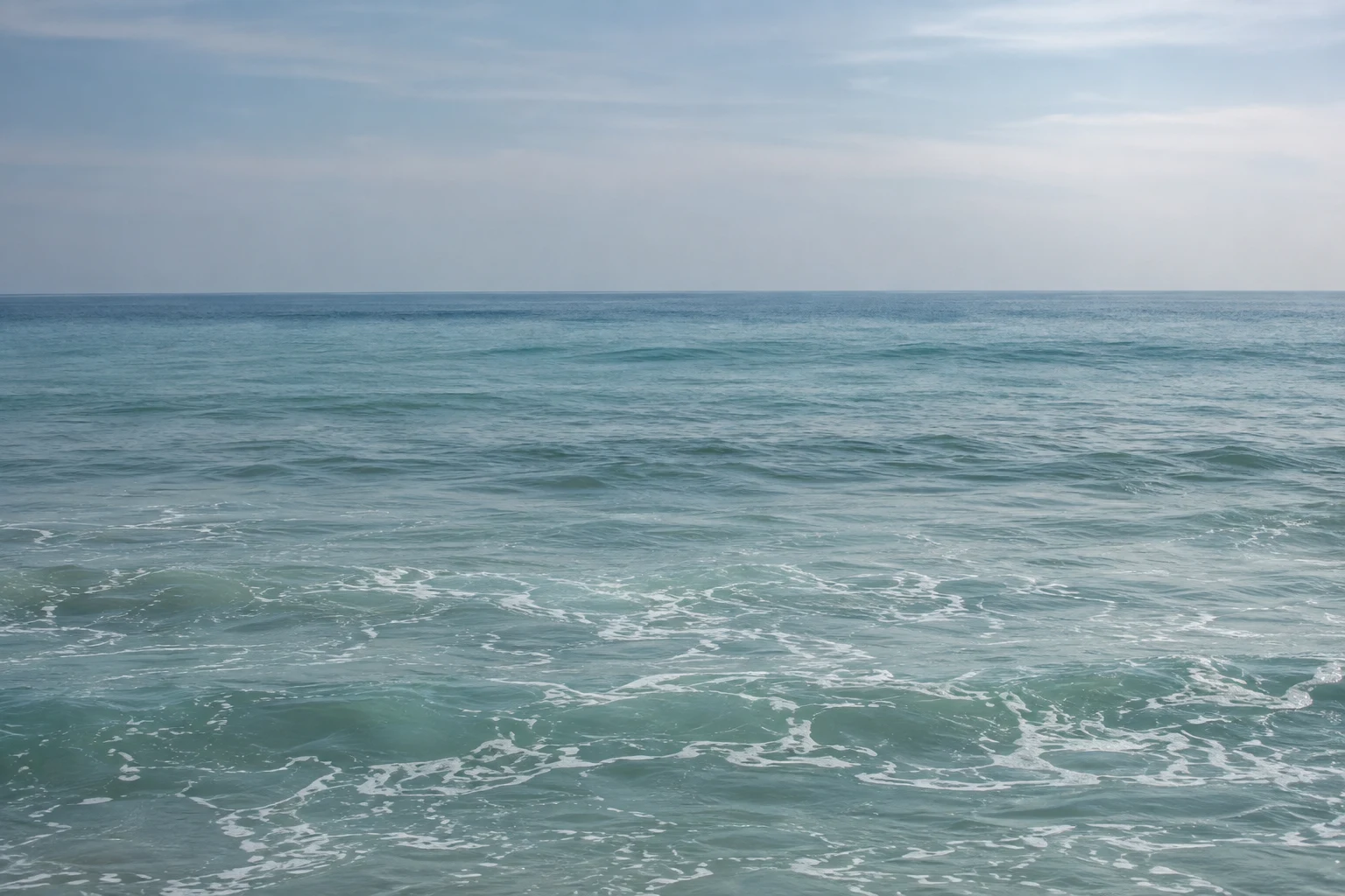 Pale teal water and fine foam soften into blue distance under a hazy sky.