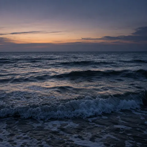 Deep navy waves hold the last soft peach light beneath a violet-gray evening sky.