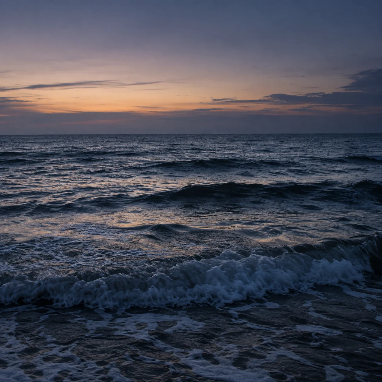 Deep navy waves hold the last soft peach light beneath a violet-gray evening sky.