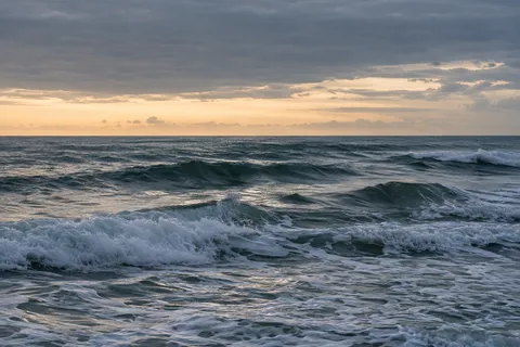 An ivory strip of sky glows below dark cloud above layered green-gray surf.