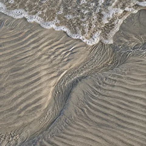 A curving shallow channel cuts through rippled pale sand toward a fine fringe of foam.