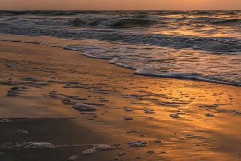 Bronze evening light reflects across wet sand and broken foam below a receding wave.