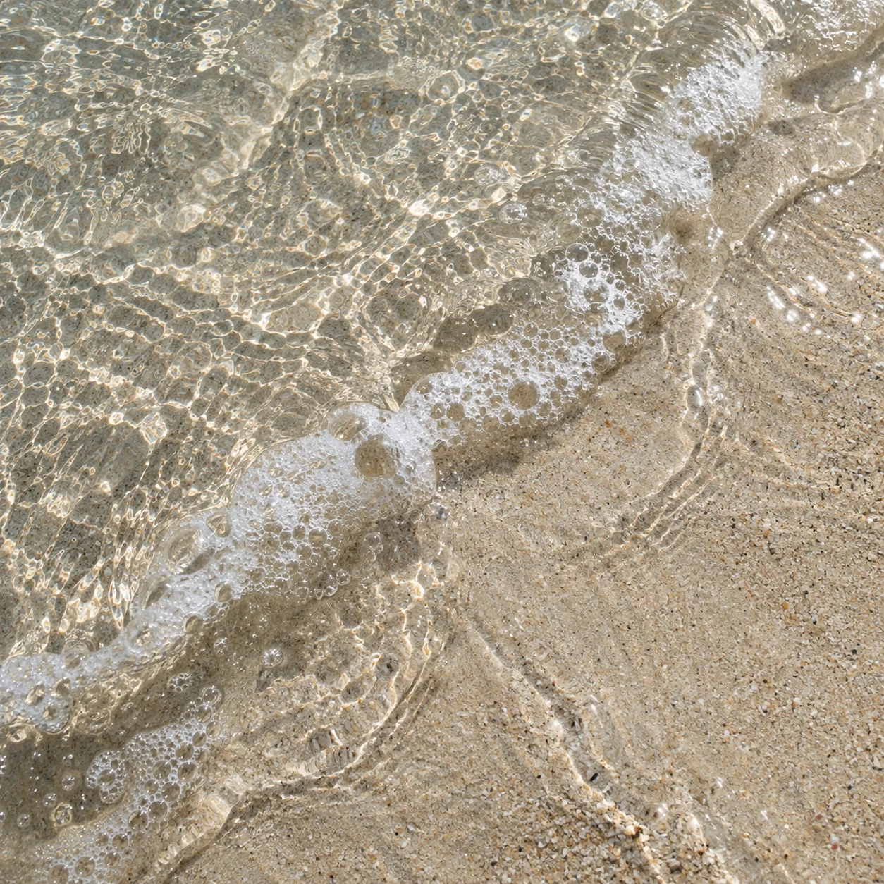 Clear water slides across pale granular sand beside a small diagonal line of bubbles.